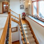 A well-lit corridor with a railing, wooden stairs leading down, and decorative items along a ledge under the windows. Listed by Andi Dyer.