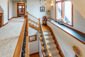 A well-lit corridor with a railing, wooden stairs leading down, and decorative items along a ledge under the windows. Listed by Andi Dyer.