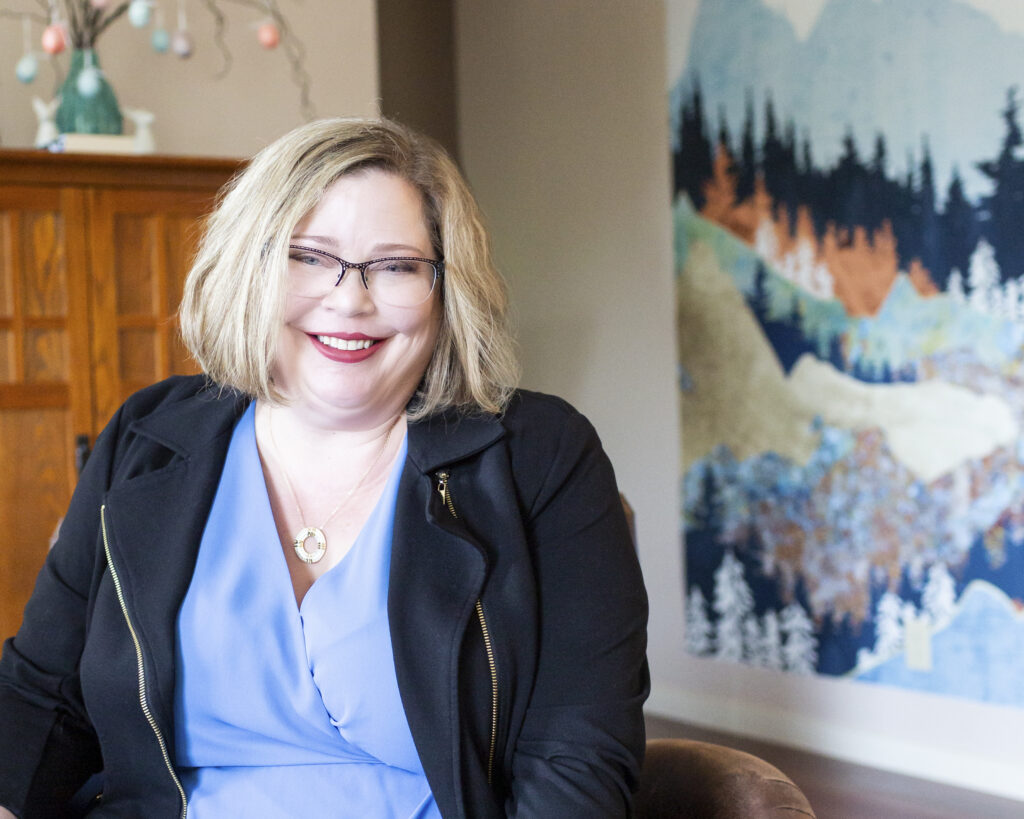 A warm and welcoming image of Andi Dyer sitting comfortably indoors. Andi is smiling confidently in a professional yet friendly pose, wearing a light blue blouse and a black jacket. Her relaxed demeanor is complemented by her stylish glasses and blonde bob haircut. The background features a cozy room setting with a wooden cabinet, a vase with decor, and an artistic wall hanging depicting a scenic landscape, adding a personal and inviting touch. This photo conveys a sense of approachability, professionalism, and warmth—qualities ideal for building trust and connection with clients.