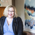 A warm and welcoming image of Andi Dyer sitting comfortably indoors. Andi is smiling confidently in a professional yet friendly pose, wearing a light blue blouse and a black jacket. Her relaxed demeanor is complemented by her stylish glasses and blonde bob haircut. The background features a cozy room setting with a wooden cabinet, a vase with decor, and an artistic wall hanging depicting a scenic landscape, adding a personal and inviting touch. This photo conveys a sense of approachability, professionalism, and warmth—qualities ideal for building trust and connection with clients.